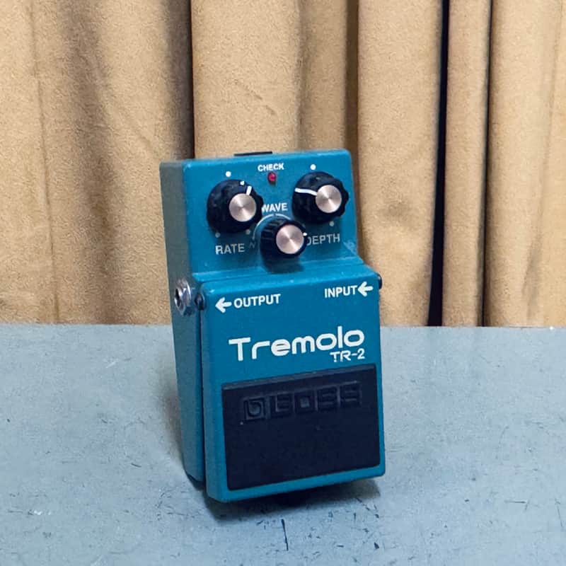 Black box and teal tremolo effect pedal on gray carpet.