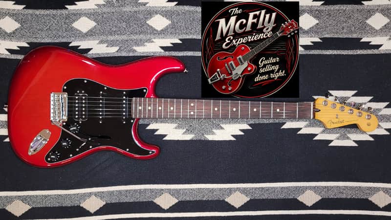 Red electric guitar with black pickguard, displayed against an American flag backdrop.