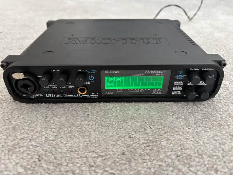 Black audio mixer with knobs, screen, and attached cables on a light surface.