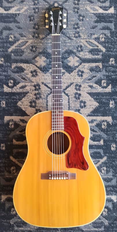Acoustic guitar with brown and red inlay, standing upright against a white wall.