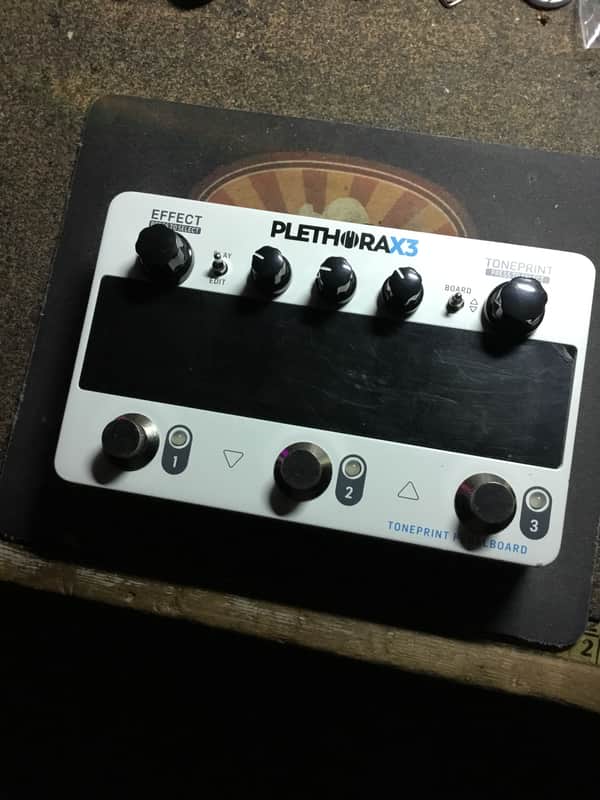 White electronic pedalboard with knobs and screen, next to open box and cables.