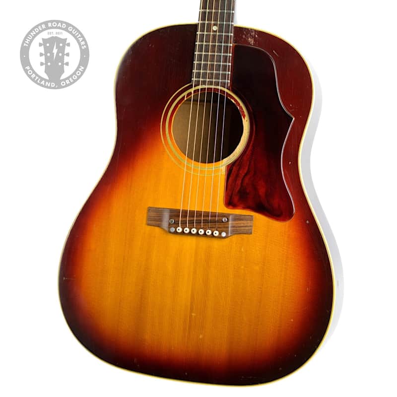 Acoustic guitar, sunburst finish, displayed vertically on a wooden floor.