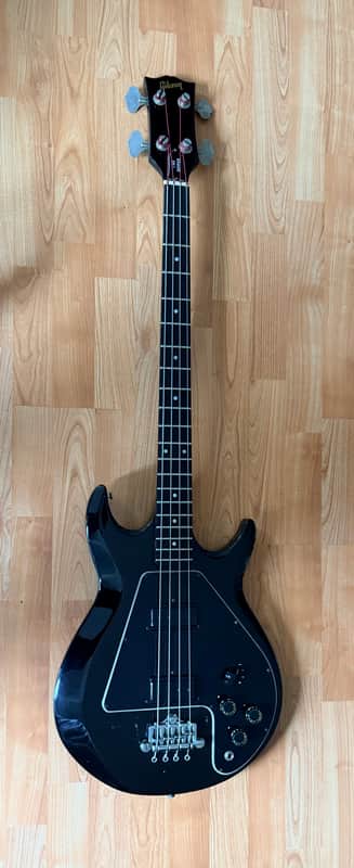 Black electric guitar, standing upright, against a light wood floor.