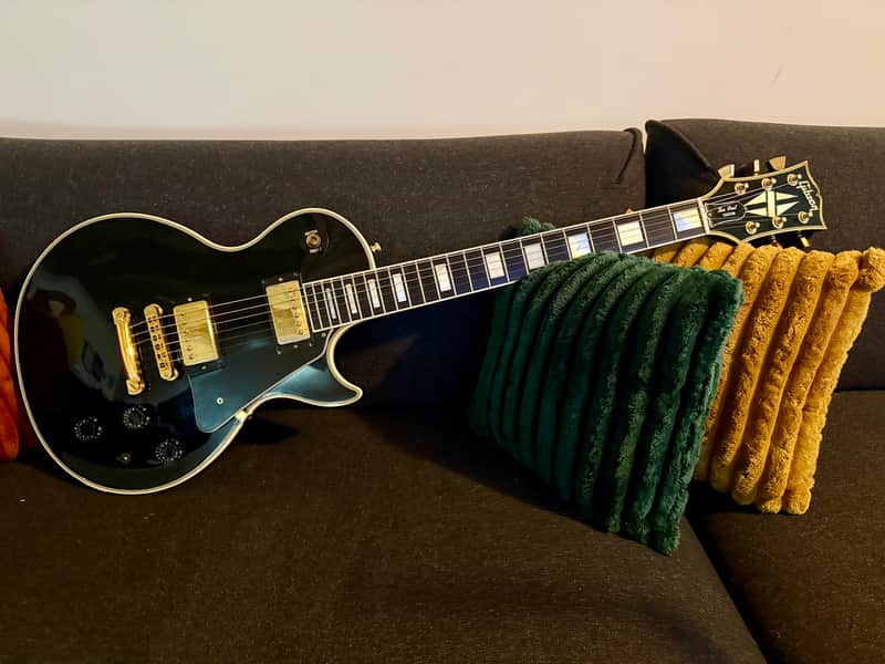 Black electric guitar with gold hardware, brown case, and accessories on wood floor.