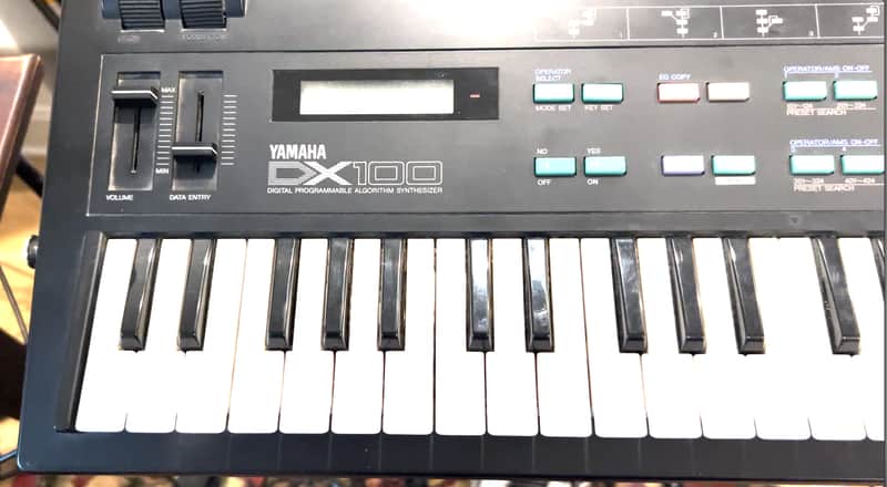 Black Yamaha DX100 keyboard resting on brown leather sofa.