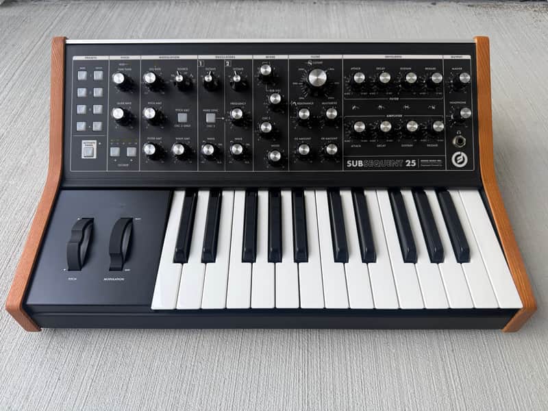 Black synthesizer keyboard with knobs and sliders, labeled "SUBSEQUENT 25.