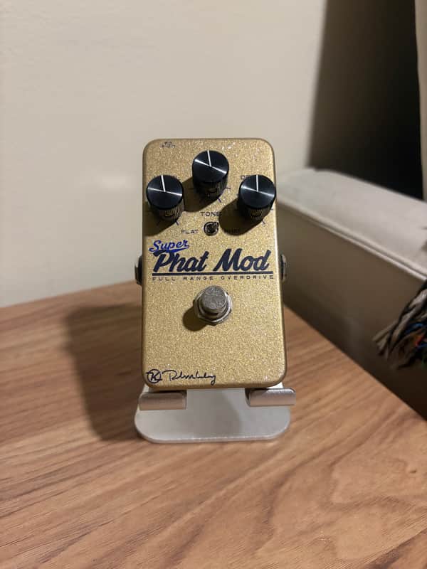 Gold overdrive pedal with knobs, labeled "Super Phat Mod," resting on white packaging.
